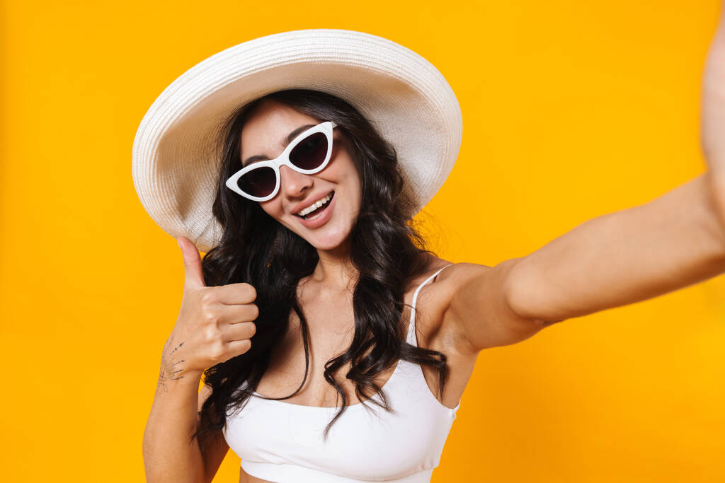 Imagen de la mujer alegre en traje de baño y sombrero gesto thunb hacia arriba mientras toma foto selfie aislado sobre la pared amarilla
 - Foto, imagen