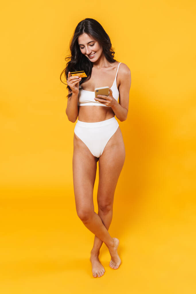 Immagine di una donna sorridente in costume da bagno con carta di credito e con il cellulare isolato sulla parete gialla - Foto, immagini