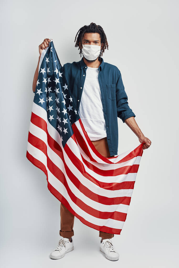 Guapo joven africano portando bandera americana con máscara facial médica y mirando a la cámara mientras está de pie sobre fondo gris
 - Foto, imagen
