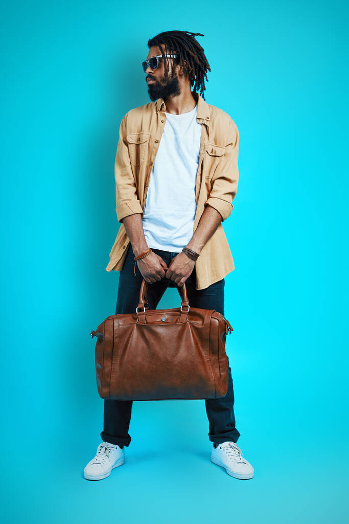Piena lunghezza di giovane uomo africano in abbigliamento casual portando borsa in pelle e guardando altrove mentre in piedi sullo sfondo blu          - Foto, immagini