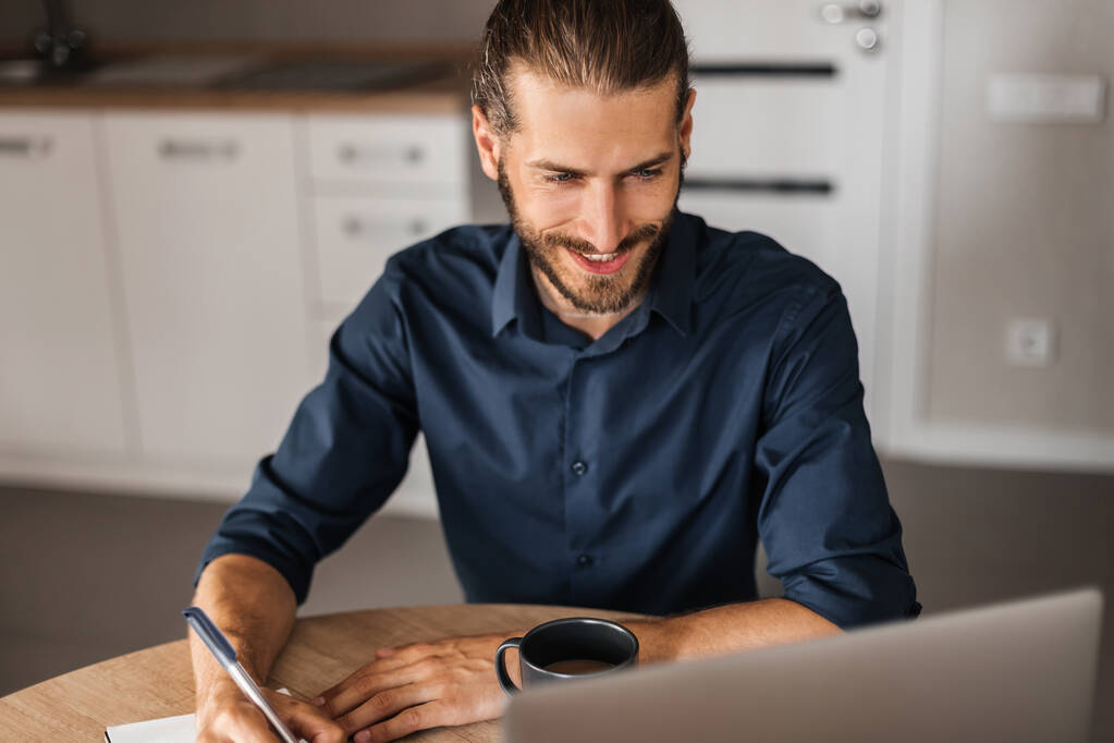 Ein Mann lächelt, arbeitet aus der Ferne am Computer. Ein junger Mann schreibt Informationen in ein Notizbuch und blickt in einen Laptop. Ein Mann arbeitet zu Hause. Blick von oben - Foto, Bild
