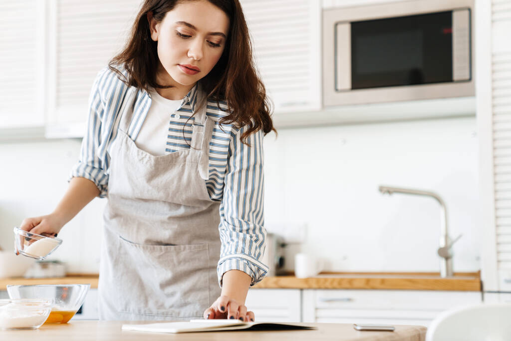 現代的なキッチンでパイを調理しながら、エプロン読書レシピを身に着けている焦点を当てた若い女性のイメージ - 写真・画像