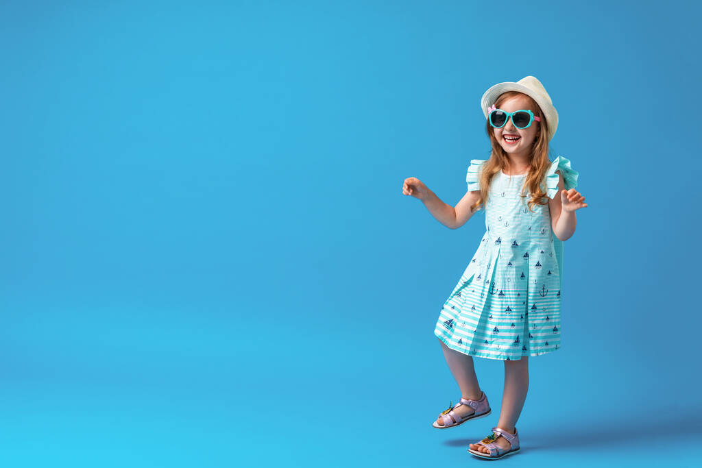 roztomilá holčička ve věku 4 let, v šatech, klobouku a slunečních brýlích, pózuje, na modrém pozadí. Dítě bude šťastné, že může začít letní prázdniny a vyrazit na výlet - Fotografie, Obrázek