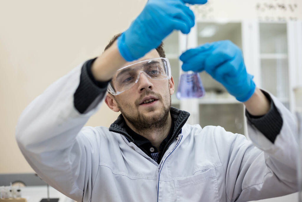 Retrato de cerca de un químico que lleva gafas de seguridad y guantes protectores, sosteniendo un frasco de líquido azul. Científico en el laboratorio
. - Foto, imagen