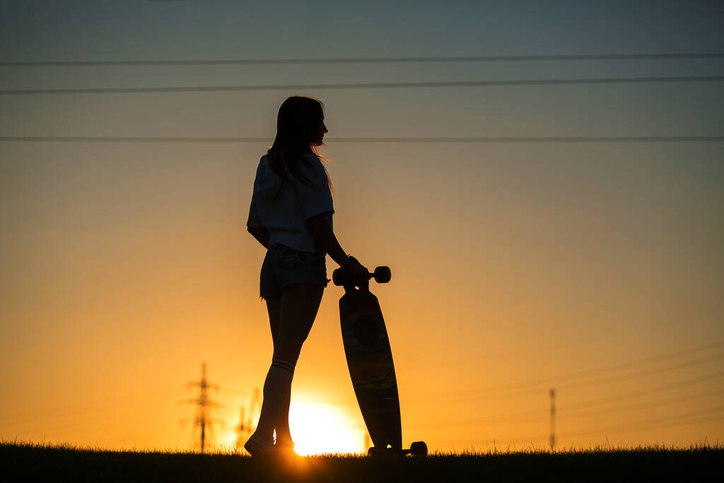 tyttö katsoo auringonlaskua pitäen longboard kädessään. - Valokuva, kuva