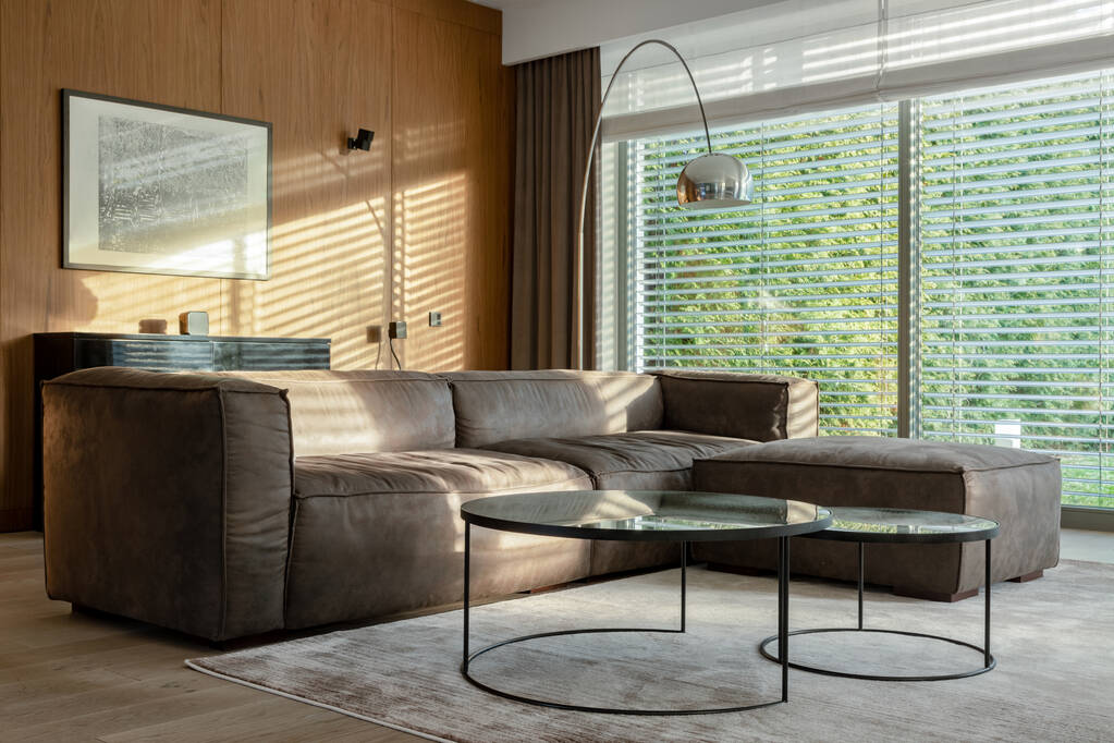 Elegancki salon z dużą sofą narożną, dwoma nowoczesnymi stolikami kawowymi i srebrną lampą z dużymi oknami z widokiem na ogród - Zdjęcie, obraz