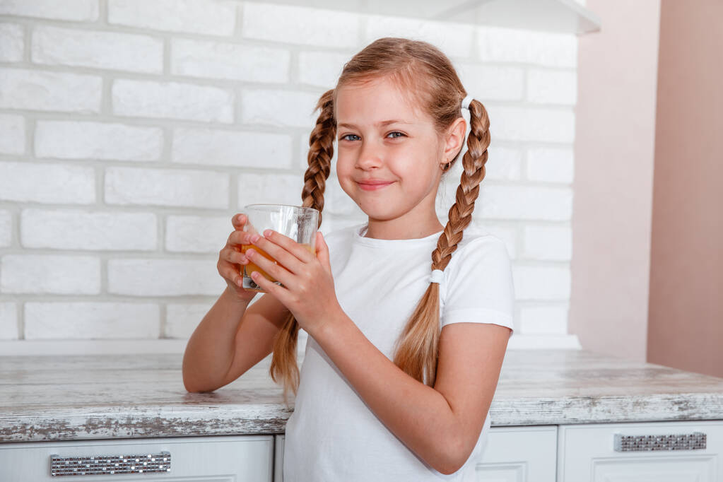 Little girl drinks fresh orange juice from a glass cup in the kitchen - Photo, Image