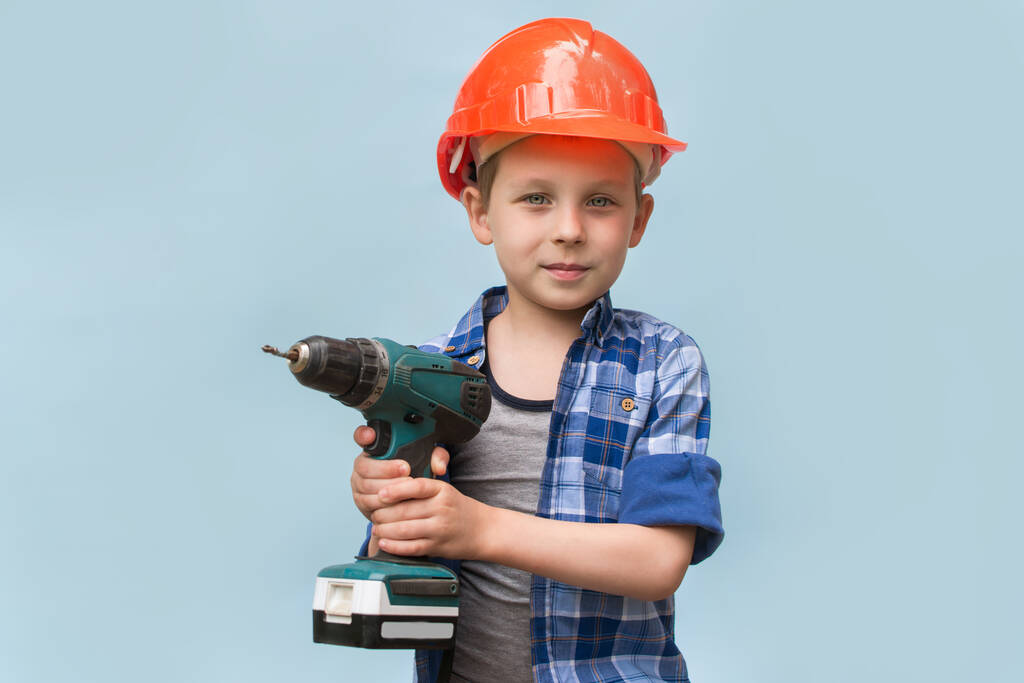 Een kleuter speelt Builder or repairman, werkend met gereedschap. Concept van kindertijd. Kleine jongen in een oranje helm of helm, op een blauwe achtergrond. Het kind droomt van een toekomstige carrière in de bouw. - Foto, afbeelding