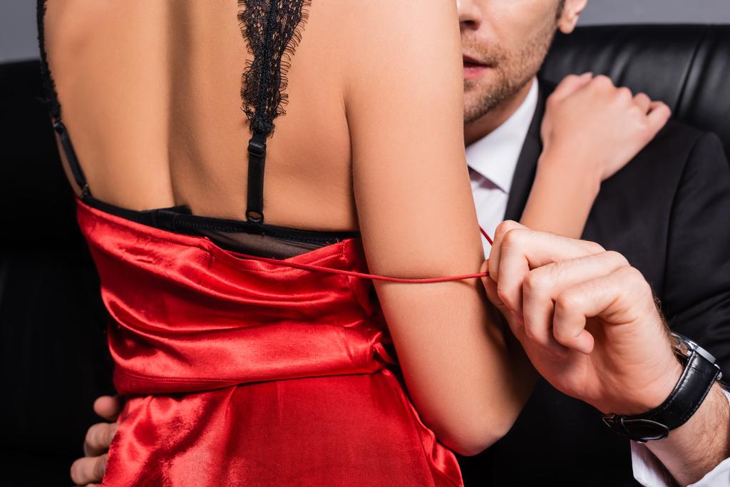 Oříznutý pohled na muže sundávat červené šaty od přítelkyně v krajkové podprsenky na gauči izolované na šedé - Fotografie, Obrázek