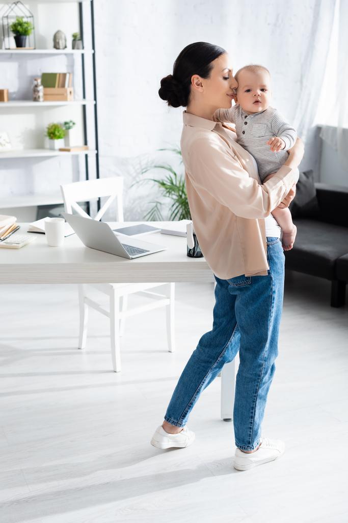 attraente madre in piedi e tenendo in braccio carino figlio neonato in pagliaccetto vicino gadget sul tavolo  - Foto, immagini