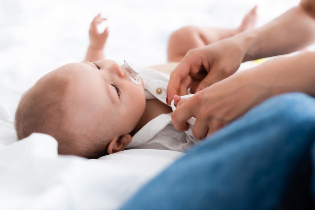 selectieve focus van moeder het dragen van baby romper op schattige zuigeling zoon met fopspeen - Foto, afbeelding