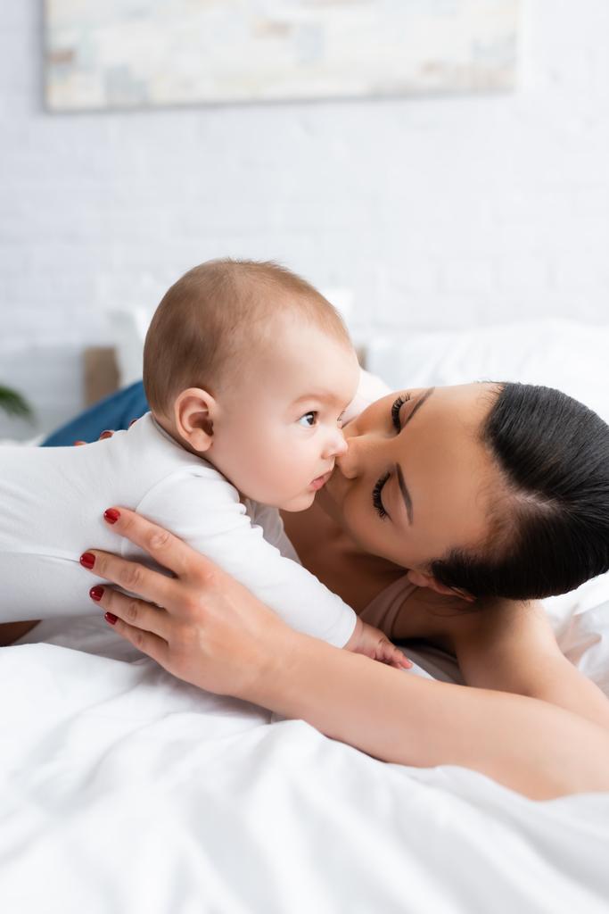 woman kissing cute infant son in baby romper lying on bed - Photo, Image