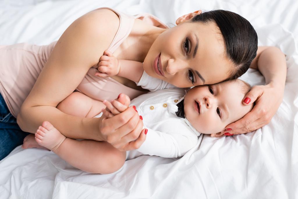 Blick über den Kopf: attraktive Mutter umarmt entzückenden Säugling im Baby-Strampler, der auf dem Bett liegt - Foto, Bild