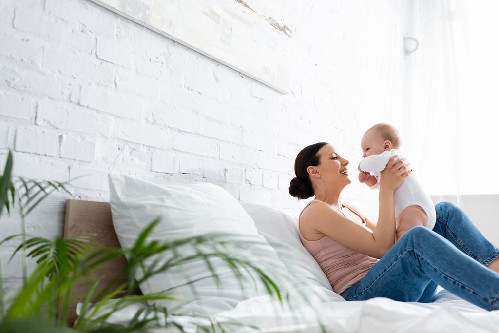 selective focus of happy mother sitting on bed and holding in arms cute infant son  - Photo, Image