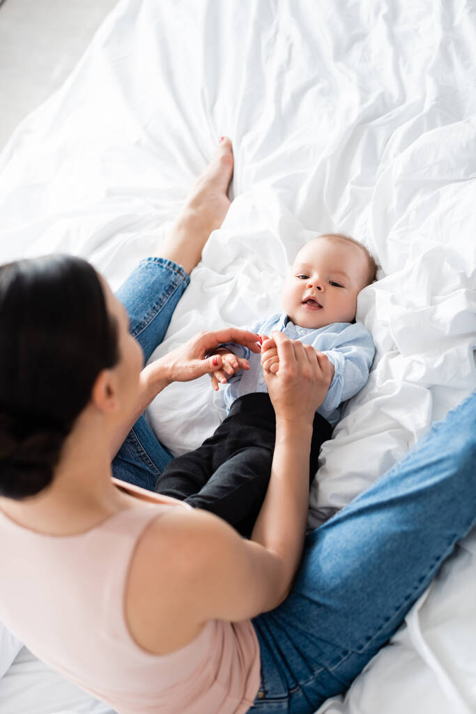 ベッドに座って可愛い赤ちゃんの息子に触れるデニムジーンズの母親のオーバーヘッドビュー  - 写真・画像