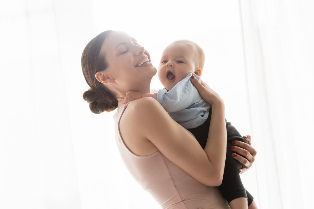 усміхнена мати з закритими очима тримає в руках немовля сина в дитячому одязі
  - Фото, зображення