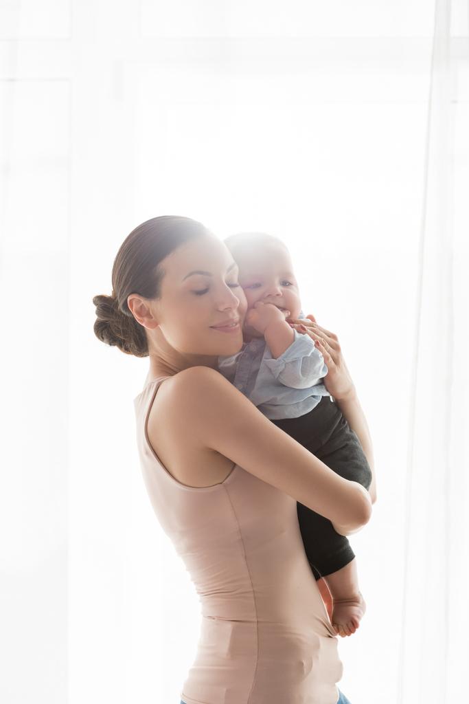 glimlachende moeder met gesloten ogen houden in armen schattige baby zoon in baby kleding  - Foto, afbeelding