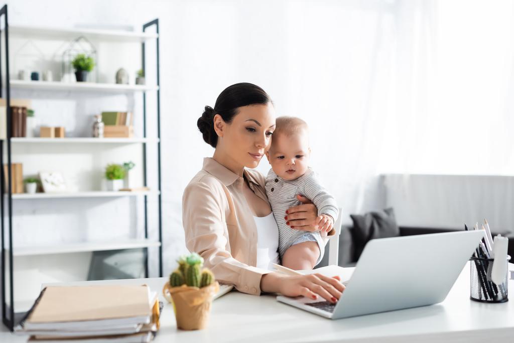 selective focus of attractive mother holding in arms baby boy and working from home - Photo, Image