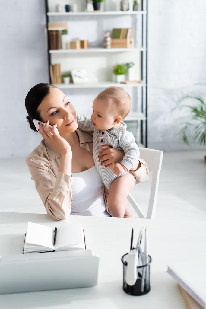 selective focus of attractive freelancer holding in arms infant son and talking on smartphone  - Photo, Image