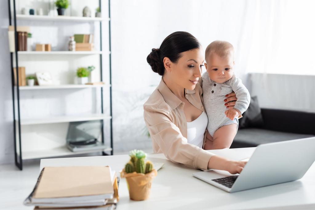 enfoque selectivo de madre atractiva sosteniendo en brazos bebé niño y el uso de ordenador portátil
 - Foto, Imagen