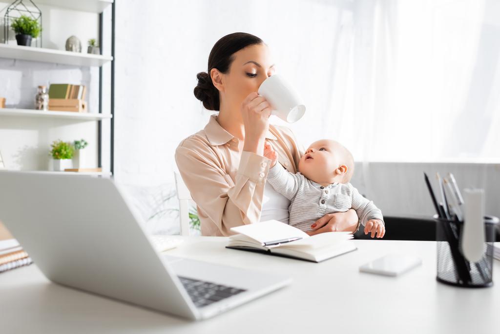 selective focus of freelancer drinking near cute infant son and gadgets  - Photo, Image