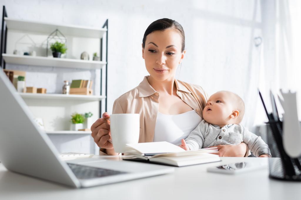 enfoque selectivo de la madre sosteniendo la taza cerca de lindo hijo pequeño mientras mira el ordenador portátil
  - Foto, imagen