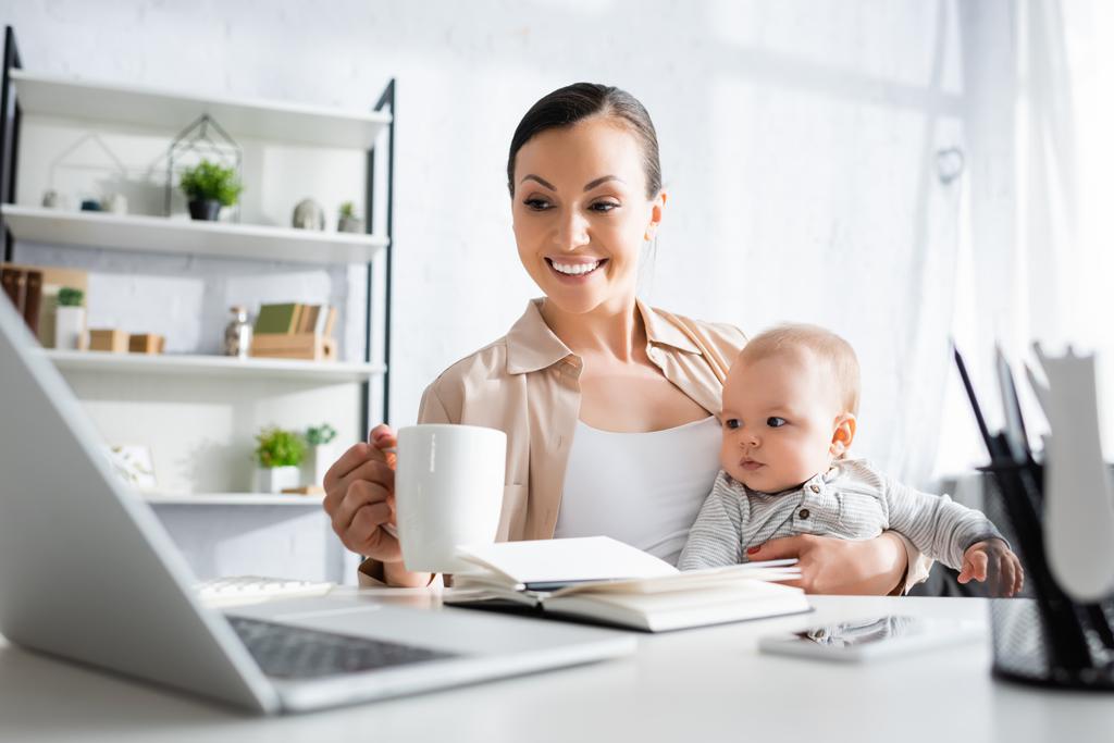 enfoque selectivo de la madre feliz sosteniendo taza cerca lindo bebé hijo y gadgets
  - Foto, Imagen