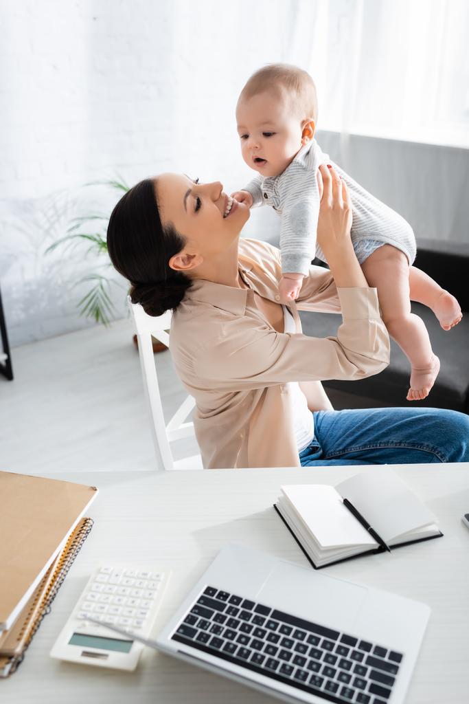 selective focus of happy mother holding in arms cute infant son near laptop  - Photo, Image