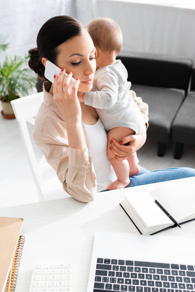привлекательная мать держит в руках младенца сына и разговаривает на смартфоне рядом с ноутбуком
  - Фото, изображение