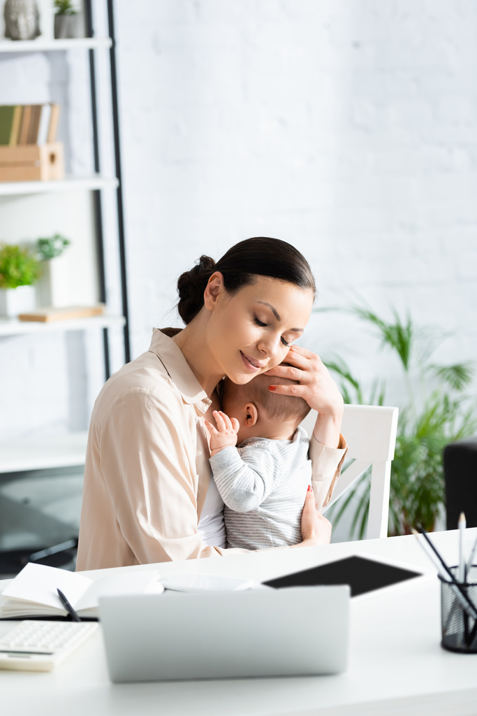 mãe com olhos fechados abraçando bebê menino perto de gadgets na mesa
  - Foto, Imagem