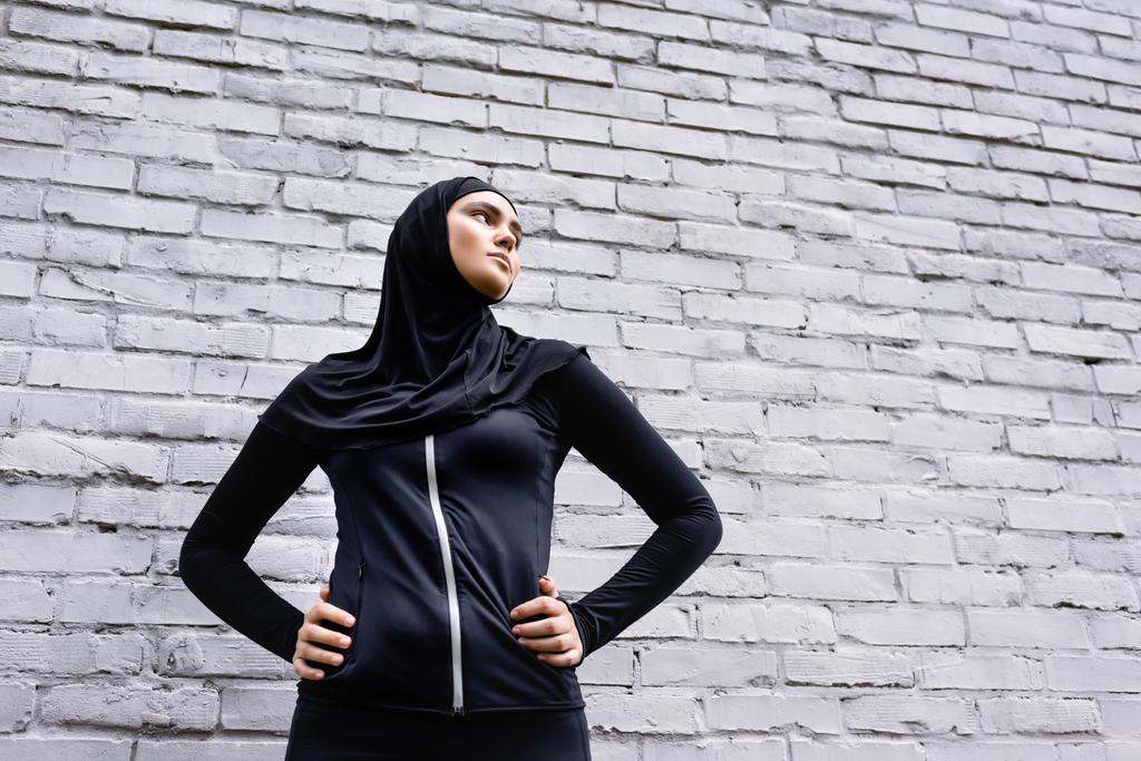 низький кут зору мусульманської дівчини в хіджабі, що стоїть руками на стегнах біля цегляної стіни
  - Фото, зображення