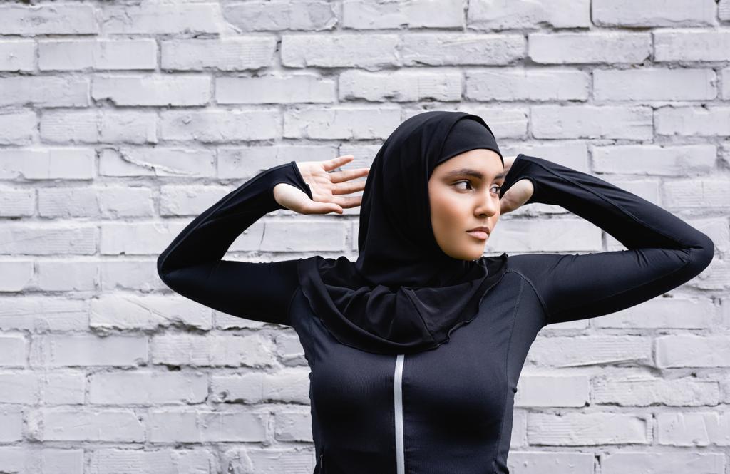 ελκυστική μουσουλμάνα γυναίκα κοιτάζοντας μακριά, ενώ η άσκηση κοντά στον τοίχο τούβλο  - Φωτογραφία, εικόνα