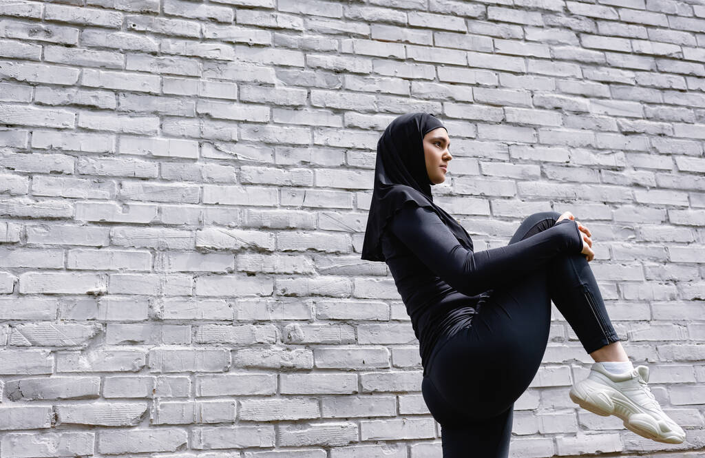 レンガの壁の近くで運動する魅力的なイスラムスポーツ女性の低角度ビュー  - 写真・画像