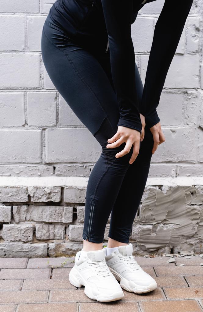 vista cortada de esportista tocando joelhos durante o exercício perto da parede de tijolo
  - Foto, Imagem