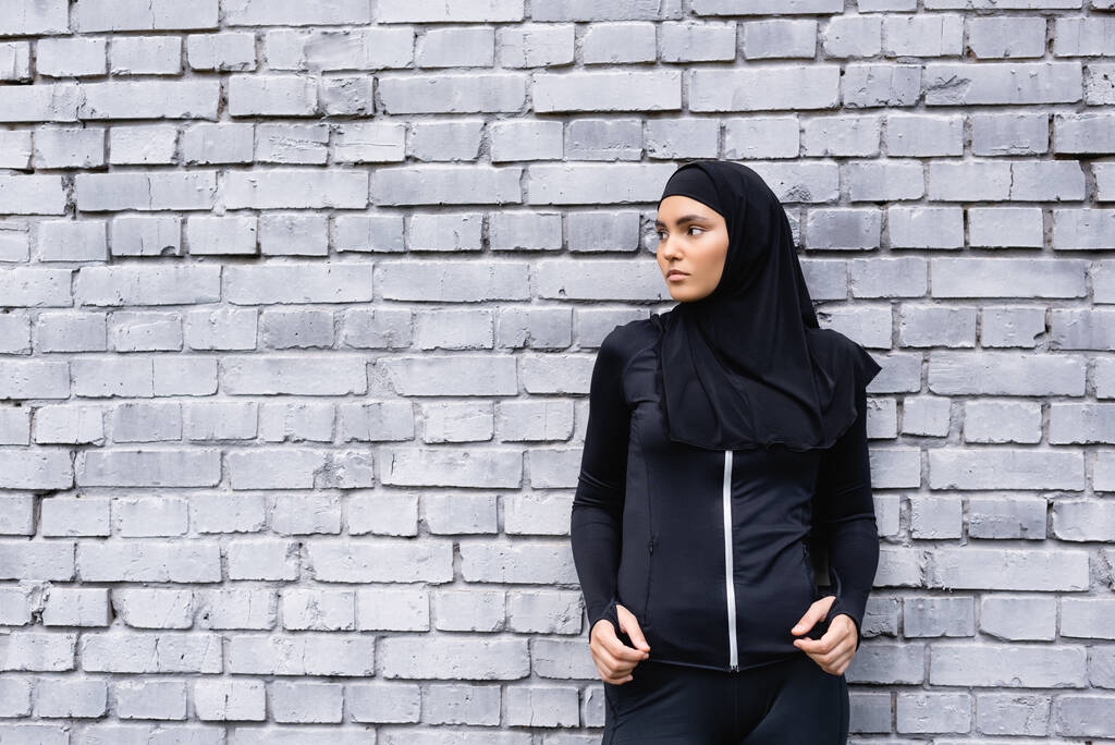 νεαρή αθλήτρια μουσουλμάνα με χιτζάμπ στέκεται κοντά σε τοίχο από τούβλα και κοιτάζει μακριά - Φωτογραφία, εικόνα