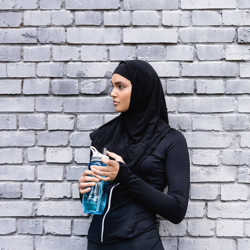 mladá muslimská sportovkyně v hidžábu drží sportovní láhev s vodou v blízkosti cihlové zdi  - Fotografie, Obrázek