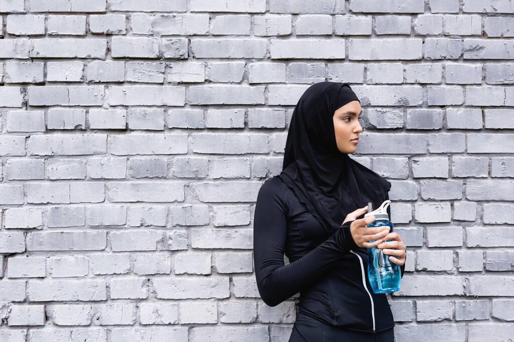młoda muzułmanka w czarnym hidżabie trzymająca butelkę sportową z wodą na zewnątrz  - Zdjęcie, obraz