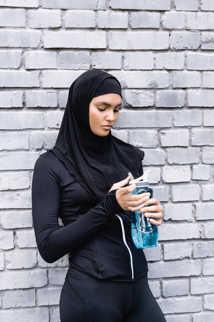 tesettürlü genç müslüman sporcu kadın dışarıda suyla birlikte spor şişesine bakıyor.  - Fotoğraf, Görsel