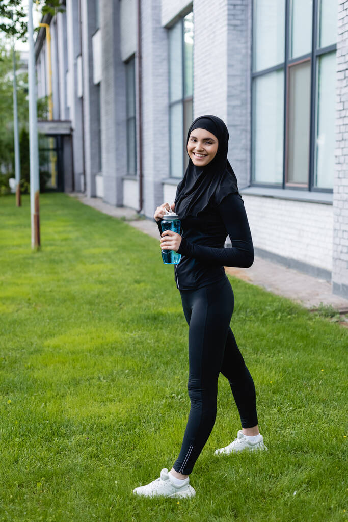 χαρούμενη αθλήτρια μουσουλμάνα στο χιτζάμπ κρατώντας αθλητικό μπουκάλι με νερό ενώ στέκεται στο πράσινο γρασίδι  - Φωτογραφία, εικόνα