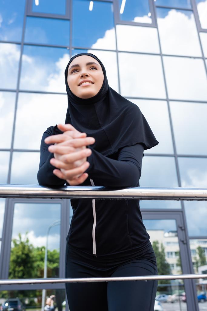 messa a fuoco selettiva di felice donna musulmana in hijab in piedi con le mani serrate vicino edificio moderno  - Foto, immagini