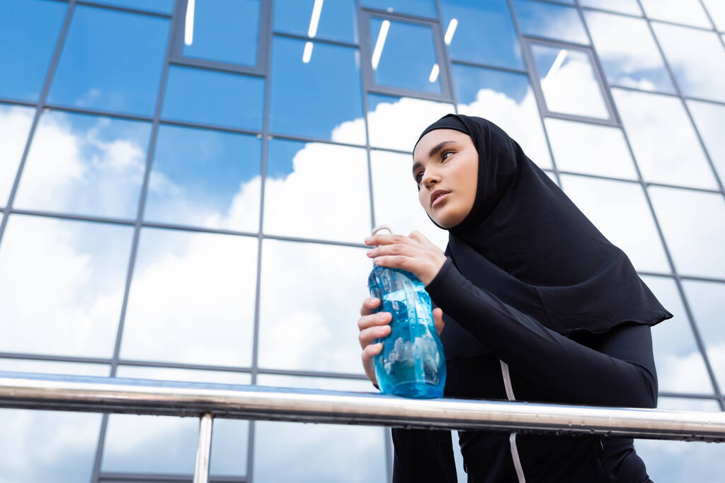 niski kąt widzenia muzułmanki w hidżabie trzymając butelkę sportową w pobliżu nowoczesnego budynku  - Zdjęcie, obraz