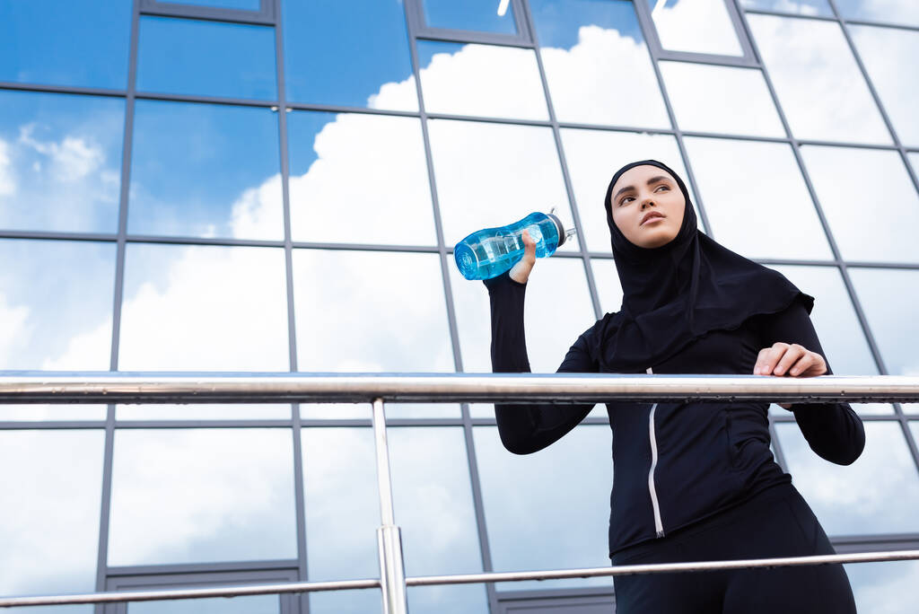 vista de ángulo bajo de la joven mujer musulmana en hijab celebración de botella deportiva cerca de edificio moderno
  - Foto, Imagen