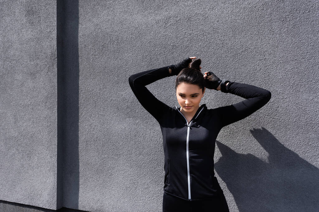 attractive sportswoman touching hair while standing near concrete wall - Photo, Image