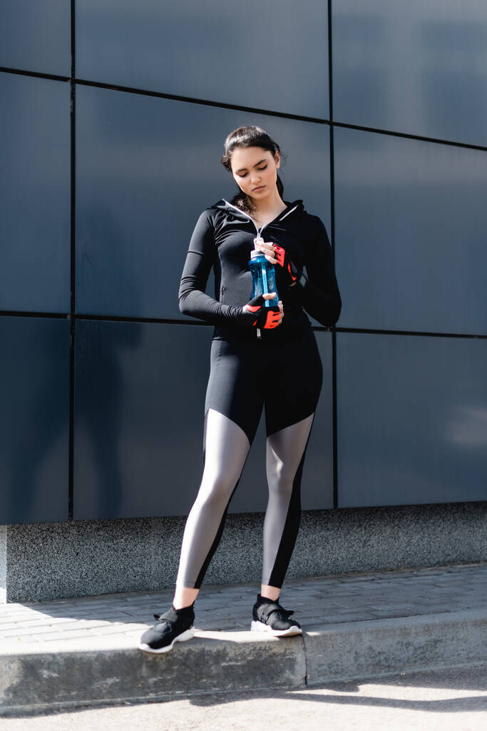 junge Frau in schwarzer Sportbekleidung hält Sportflasche mit Wasser in der Hand  - Foto, Bild