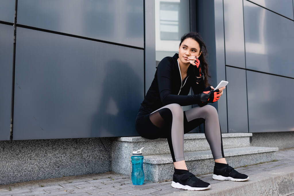 young sportswoman sitting near sports bottle and holding smartphone  - Photo, Image