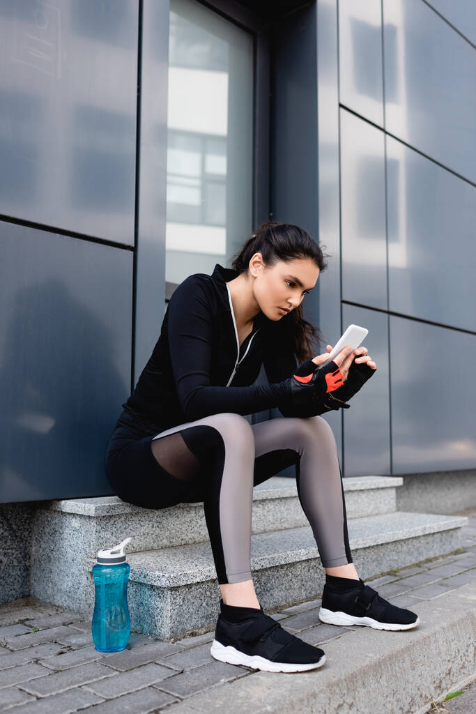 молодая спортсменка сидит рядом со спортивной бутылкой и пользуется смартфоном
  - Фото, изображение