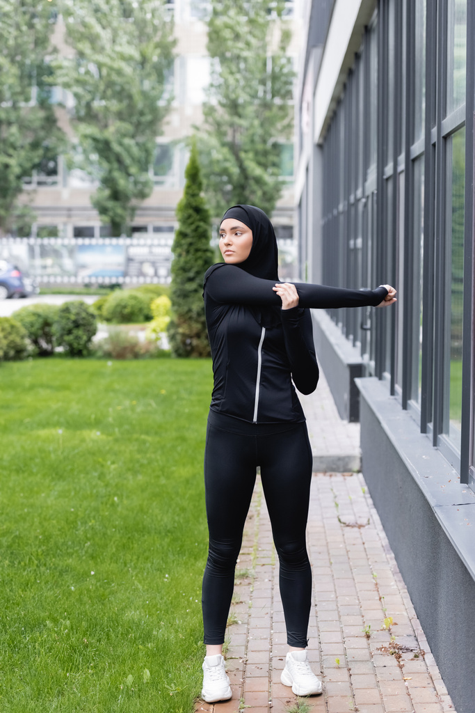 arabialainen nainen hijabissa ja urheiluvaatteet toimivat lähellä rakennusta ja vihreää ruohoa  - Valokuva, kuva