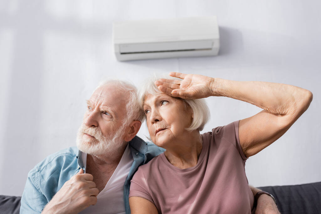 Tired senior couple looking away while feeling hot on couch under air conditioner  - Photo, Image