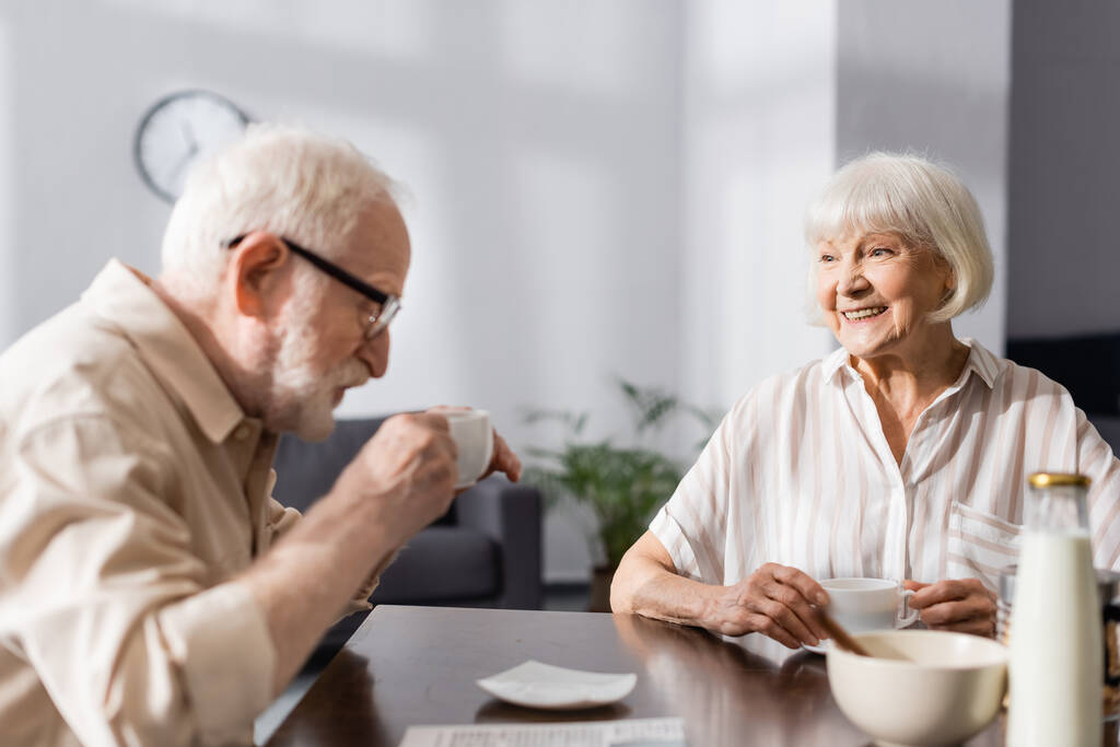 Επιλεκτική εστίαση του χαμογελαστού ηλικιωμένη γυναίκα κοιτάζοντας το σύζυγο πίνοντας καφέ στο σπίτι  - Φωτογραφία, εικόνα