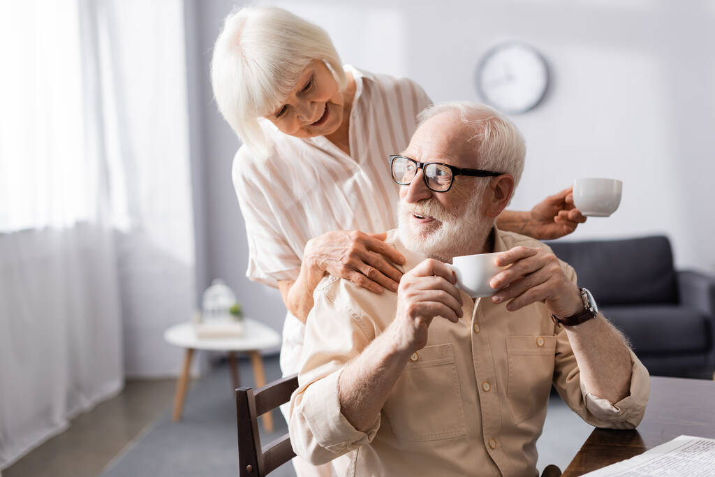 Lächelnde Seniorin hält Tasse Kaffee in der Hand und umarmt Ehemann beim Frühstück zu Hause  - Foto, Bild